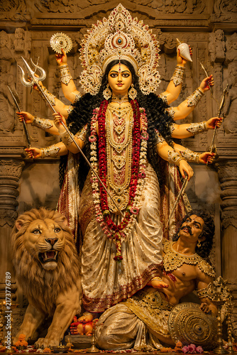 Majestic Goddess Durga idol standing with multiple arms, lion and Mahishasura, wearing elaborate crown, jewelry, and garlands inside richly carved traditional Hindu festival shrine setting.