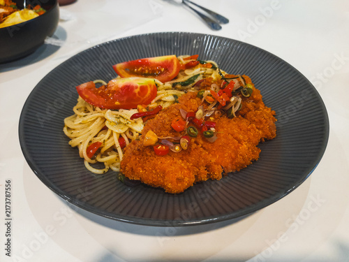 Spaghetti aglio olio served with crispy chicken katsu, sliced chili, fresh tomato, and sambal matah on a textured gray ceramic plate, combining Italian pasta with Indonesian flavors.