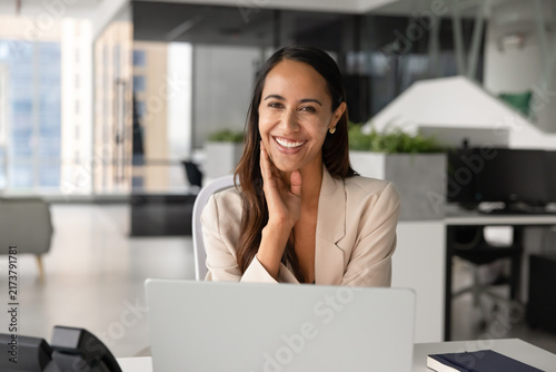 Portrait of smiling Maori businesswoman or office employee sits at workplace desk with laptop, looks at camera, represents corporate success, professionalism, career growth, business goals achievement