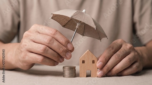 Hands Holding Small Umbrella Over Wooden House Model and Stack of Coins for Property Insurance and Financial Protection Concept