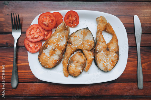 Pieces of fish fried in sesame seeds sit on a white plate next to tomato wedges. Served with a knife and fork on a brown wooden table. A perfect dish for breakfast, lunch, or dinner.
