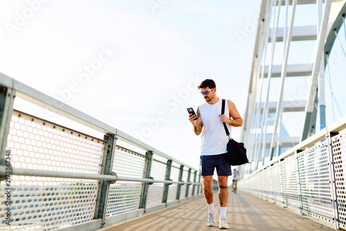 Man carrying a gym bag and checking his smartphone while walking on a modern bridge after a fitness workout