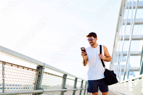 Photography Young man wearing sportswear and sunglasses walking on a modern bridge, using hi