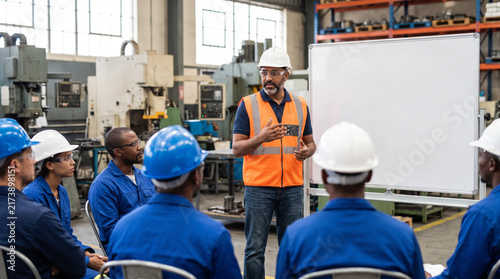 Factory safety instructor leading a training session for a group of workers in a manufacturing plant. Group of employees in hard hats attending a workplace safety briefing beside a whiteboard
