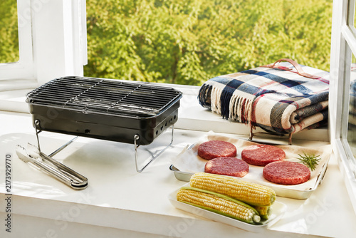 Barbecue grill, hamburger patties, corn and picnic blanket on window ledge, late summer weekend still life