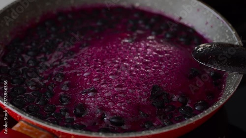 Fresh blueberries and rich purple juice boil and bubble in a red frying pan on the stove while making homemade berry jam.