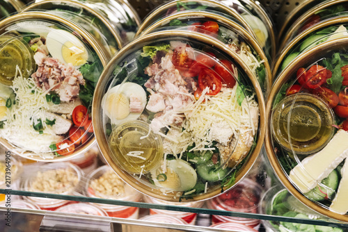 Fresh prepared salads in transparent takeaway bowls inside refrigerated display. Vegetables eggs cheese and meat form healthy food background. Ready meals for quick lunch and retail sale.