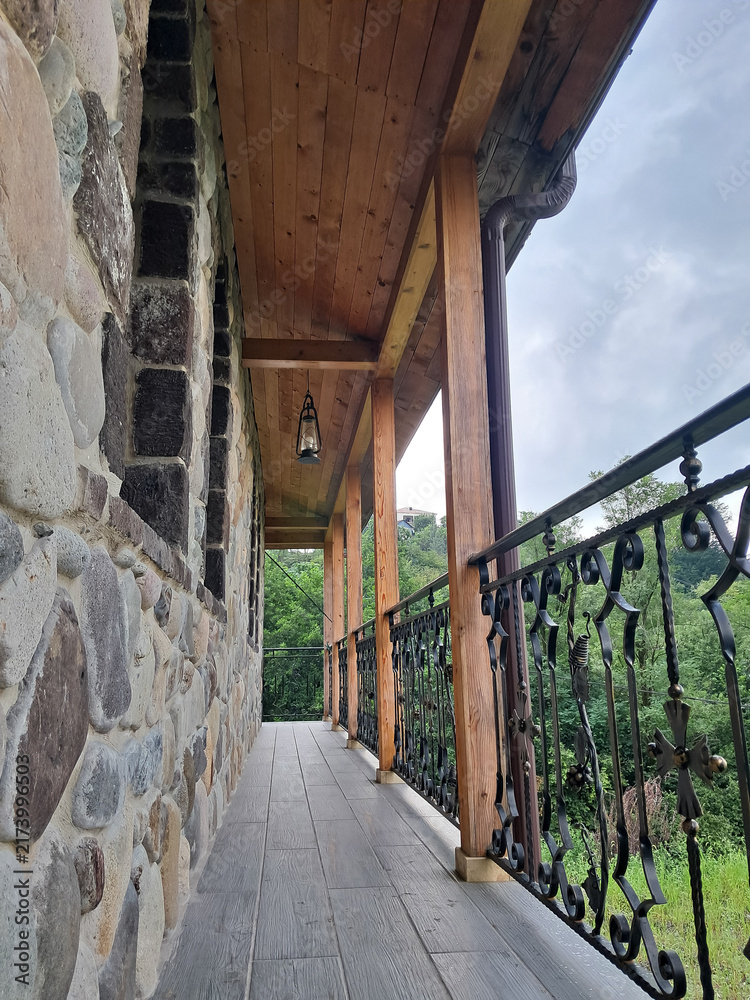 Naklejka premium Covered wooden balcony with a stone wall and ornate metal railing overlooking greenery