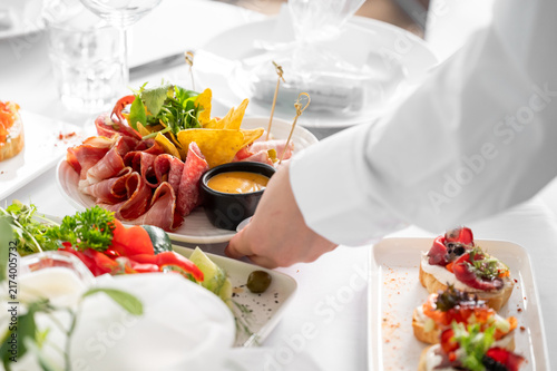 Cuadro en lienzo A waiter in a white shirt sets down a plate of appetizers, including cured meats, chips, and dip, on a restaurant table featuring bruschetta and other finger foods