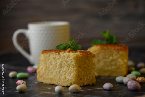 Fresh homemade sponge cake pieces with mint garnish and colorful candy dragees on a dark surface