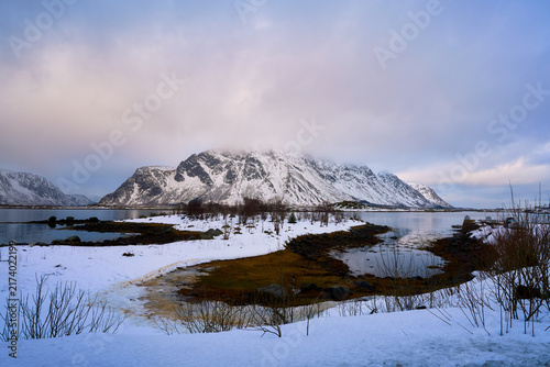 Beautiful winter landscape of Lofoten islands in Norway with grand snow-covered mountains rising from the blue ocean. Scenic high-angle viewpoint of Arctic nature under a dramatic cloudy sky in Scandi