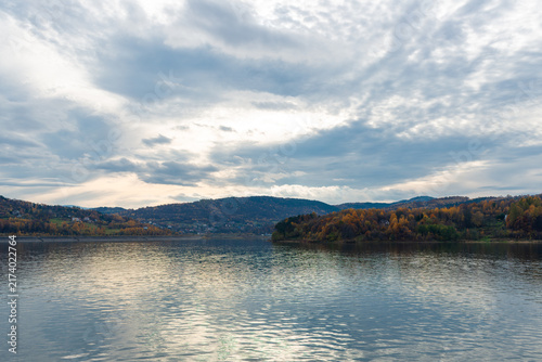 Fotografía Calm water of Jezioro Mucharskie reservoir reflects a cloudy autumn sky beneath forested Beskid hills in southern Poland