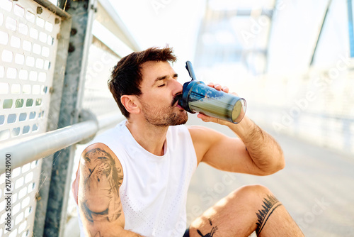 Fotografie Male athlete hydrating with a protein shake on a bridge, recovering after an out