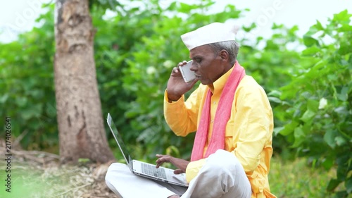 indian senior farmer talking on mobile phone while using laptop at agriculture field