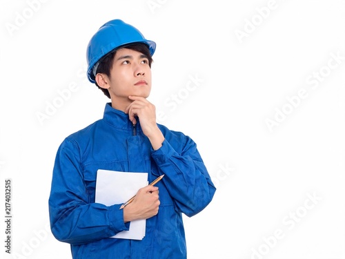 Unsure young male engineer wearing safety helmet and uniform holding note pad and pencil keeping hand on chin looking up isolated on white background
