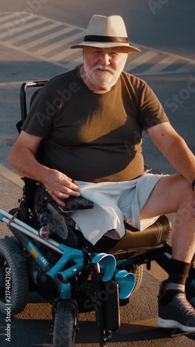 Vertical portrait of a smiling senior man in an electric wheelchair positioned on a parking space marked with the international symbol of accessibility, looking confidently at the camera in an urban s