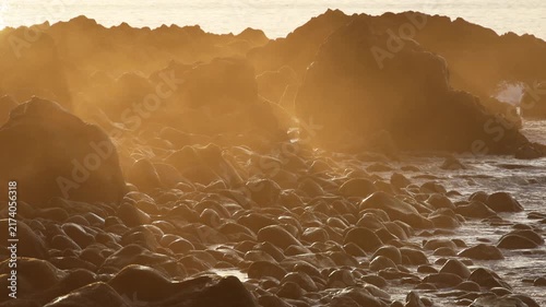 Golden sunset light shining through ocean mist over the rocky volcanic pebble coast with crashing waves in Madeira island, creating an ethereal peaceful mood. 4K video.
