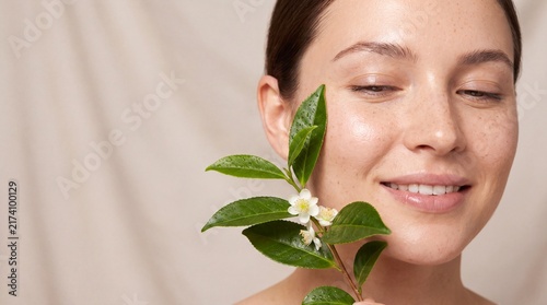 Young woman with glowing skin holding a fresh green tea branch with white flowers near her face with natural complexion posing