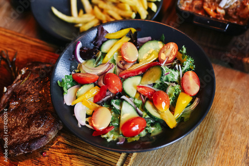 Obraz na plátne Fresh mixed vegetable salad in black bowl beside grilled steak and fries