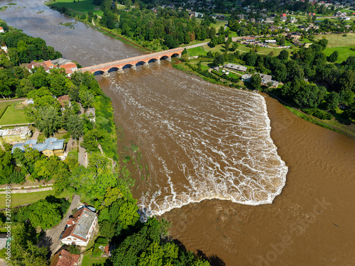 Aerial drone view of Ventas Rumba, Europe's widest waterfall in Kuldiga, Latvia, featuring the historic red brick bridge spanning across the muddy river surrounded by lush green summer landscape.