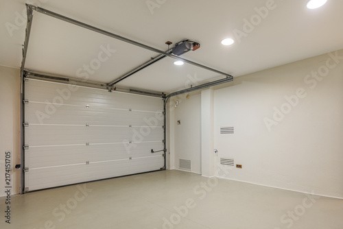 Clean empty residential garage with closed white sectional door bright overhead lighting and smooth light-colored floor
