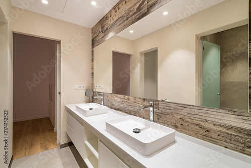 Modern bathroom interior with double countertop sinks wooden accent and large mirror under bright recessed lighting