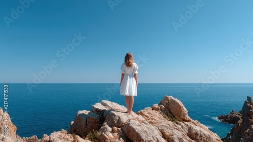 A woman stands barefoot on rocky terrain at the ocean's edge, exuding tranquility and contemplative silence against a backdrop of blue skies and calm waters.