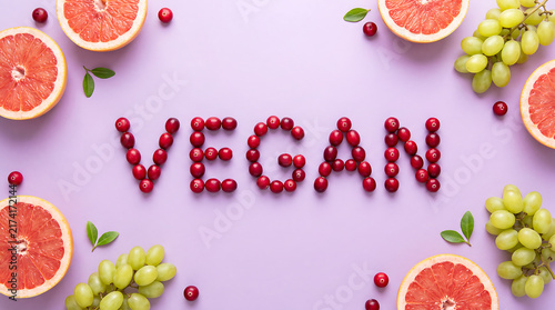 Top view flat lay of the word VEGAN spelled with fresh red cranberries surrounded by sliced pink grapefruits and green grapes on a lilac background.