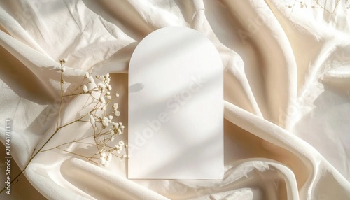 Arched paper card mockup with delicate baby breath flowers on draped beige fabric backdrop, wedding stationery invitation display