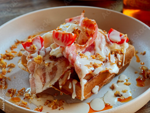 Food is presented on a plate. The dish includes bread topped with bacon, sauce, and garnishes. The setting shows a casual eating environment with good lighting