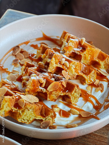 Waffles are served on a plate with drizzled caramel sauce and topped with sliced almonds. The dish is placed on a wooden table and looks appealing