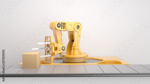Yellow robotic arm gripping a cardboard box with metal forks on a gray conveyor belt against a plain white background.