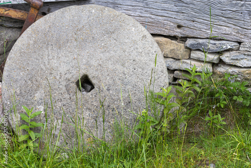 Quadro su tela a historic circular grindstone resting in green overgrowth, positioned against a rural exterior wall constructed of stacked rock and aged timber