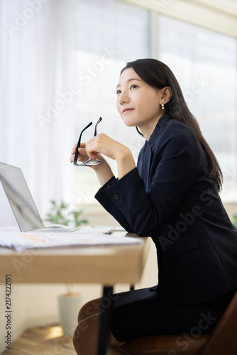 Young Japanese businesswoman holding her glasses and smiling while looking out of the window at a desk