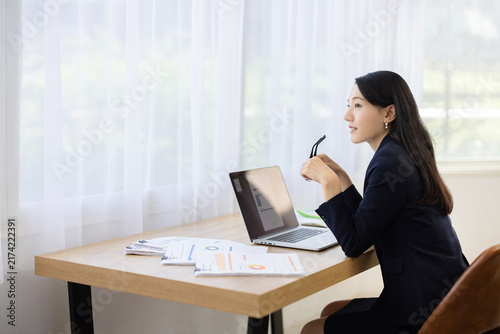 Young Japanese businesswoman holding her glasses and smiling while looking out of the window at a desk