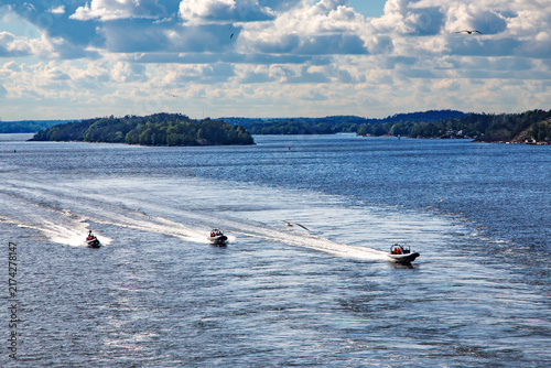A seascape featuring three high-speed RIB boats moving across the waters of a picturesque Stockholm archipelago with forested islands and cloudy skies.
