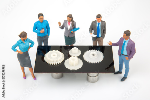 Miniature people discuss gears on a table in a bright room