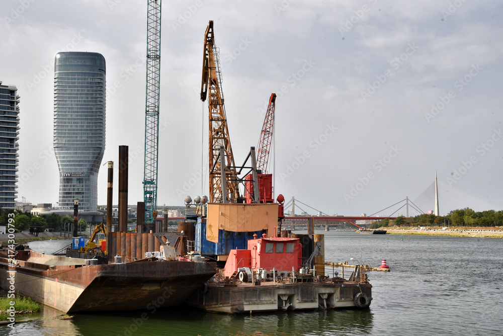 Fototapeta premium Construction Barges and Heavy Cranes on Riverfront Near Skyscraper