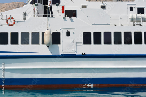A white passenger ferry ship with black tinted windows and a blue stripe floats on sea water with a large fender hanging from the side.