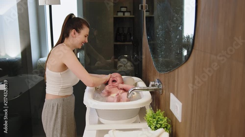 Mother gently washes crying infant in portable bathtub water splashing bathroom setting with wooden accents and modern fixtures. A mother lovingly bathes crying infant showing hygiene care and bonding