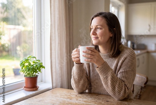 Woman holding a cup while sitting at a table by the window in a bright kitchen during the day