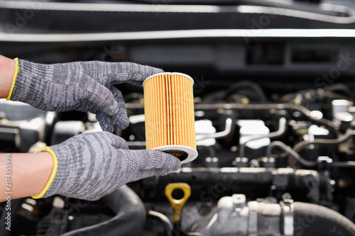 Close up of mechanic hands in protective gloves holding yellow paper oil filter element over open car engine bay, demonstrating routine automobile service and replacement