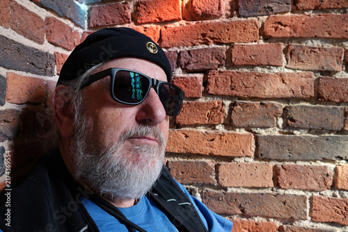 Elderly man with a beard wearing dark sunglasses and a black beanie with a small anchor.