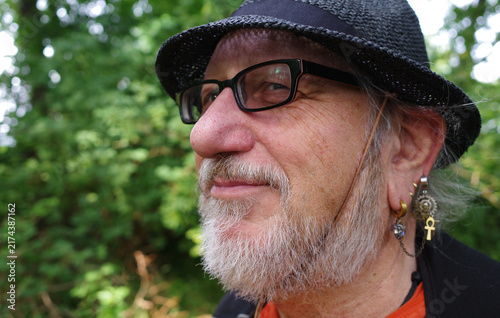 Man in profile, in his sixties, wears a textured black hat and glasses. He sports a neatly trimmed gray beard. Multiple earrings, including an ankh and other intricate designs, adorn his left ear. 