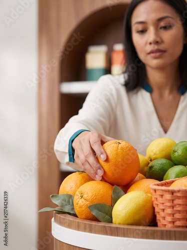 A woman in a white blouse selects a fresh orange from a wooden display shelf in a grocery store, captured in a medium shot with soft indoor lighting.