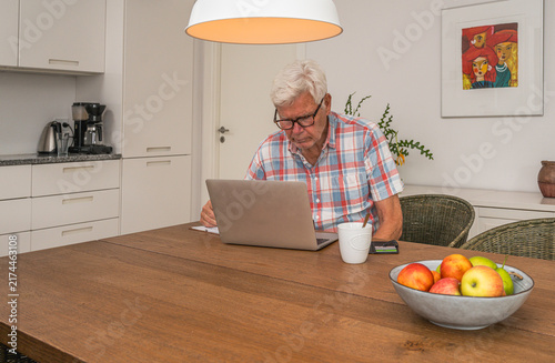 An elderly man with white hair sitting at a wooden dining table writing in a notebook
