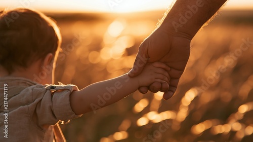 Father holding his young child's hand while walking together through a sunlit field at sunset.