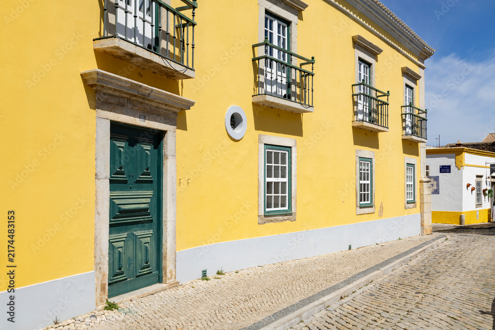 Fototapeta premium Faro Algarve Portugal 21st march 2026 Sunlit Yellow Facade With Green Door And Small Balconies On A Cobblestone Street
