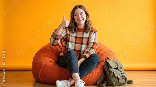 Happy young female student sitting in a bean bag chair showing thumbs up on yellow background