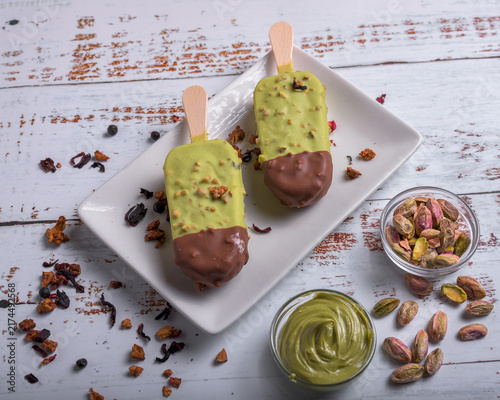 Artisanal pistachio ice cream bars dipped in chocolate served on a white plate with pistachio cream, shelled nuts, and star anise on wooden background